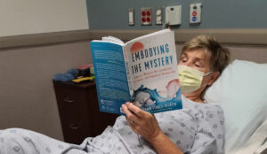 Sally Helgesen in a hospital bed reading "Embodying the Mystery" by Richard Strozzi-Heckler